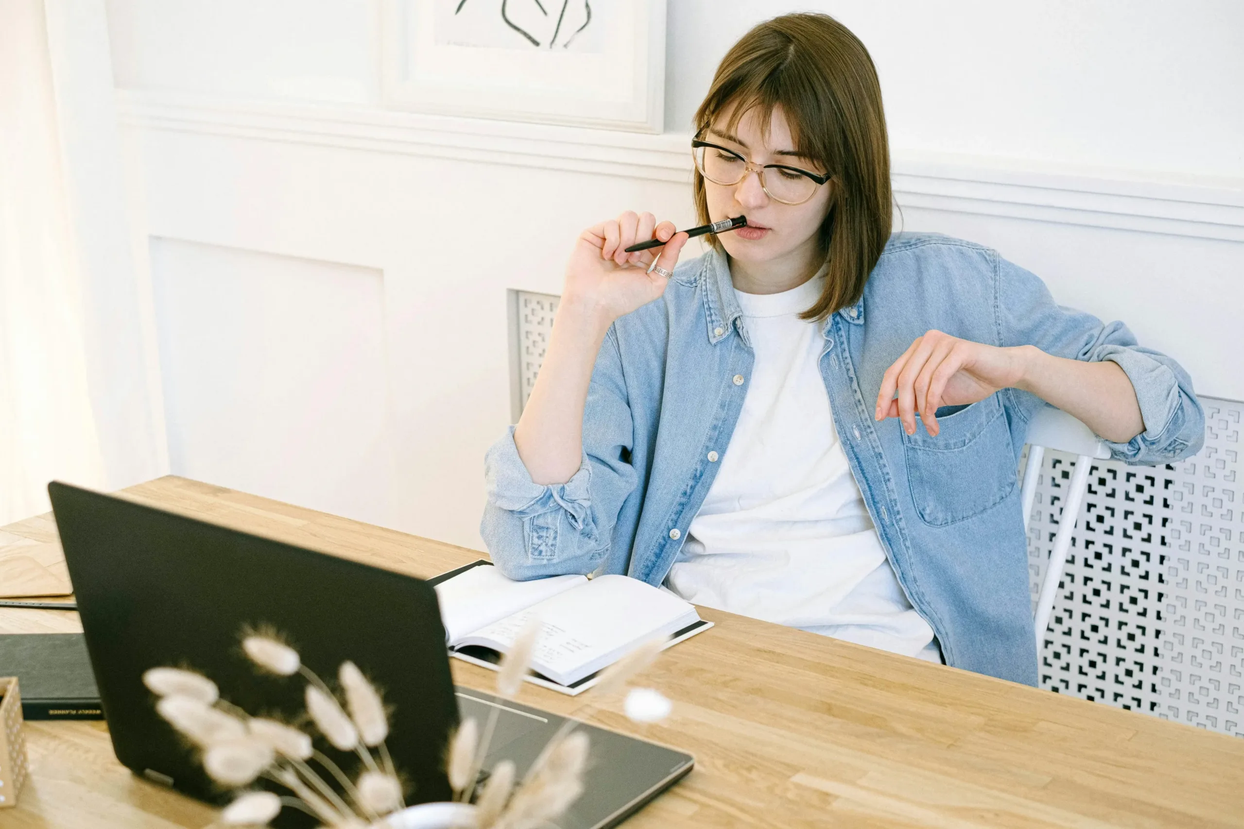 woman holding pen to her lips on desk with laptop and notebook